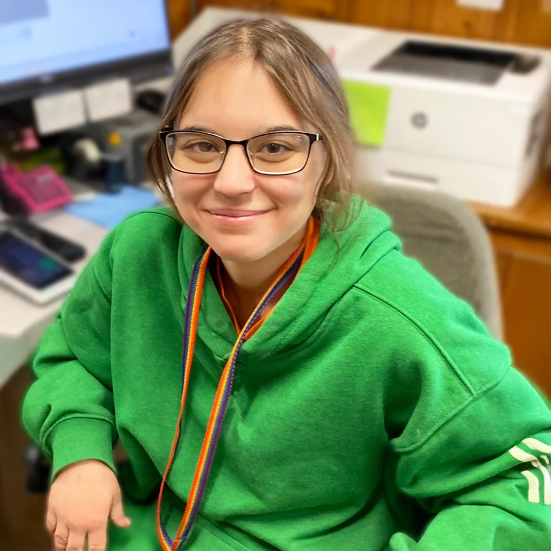 Angel Miles – Young woman wearing glasses and a green hoodie sitting at a desk and smiling at the camera.