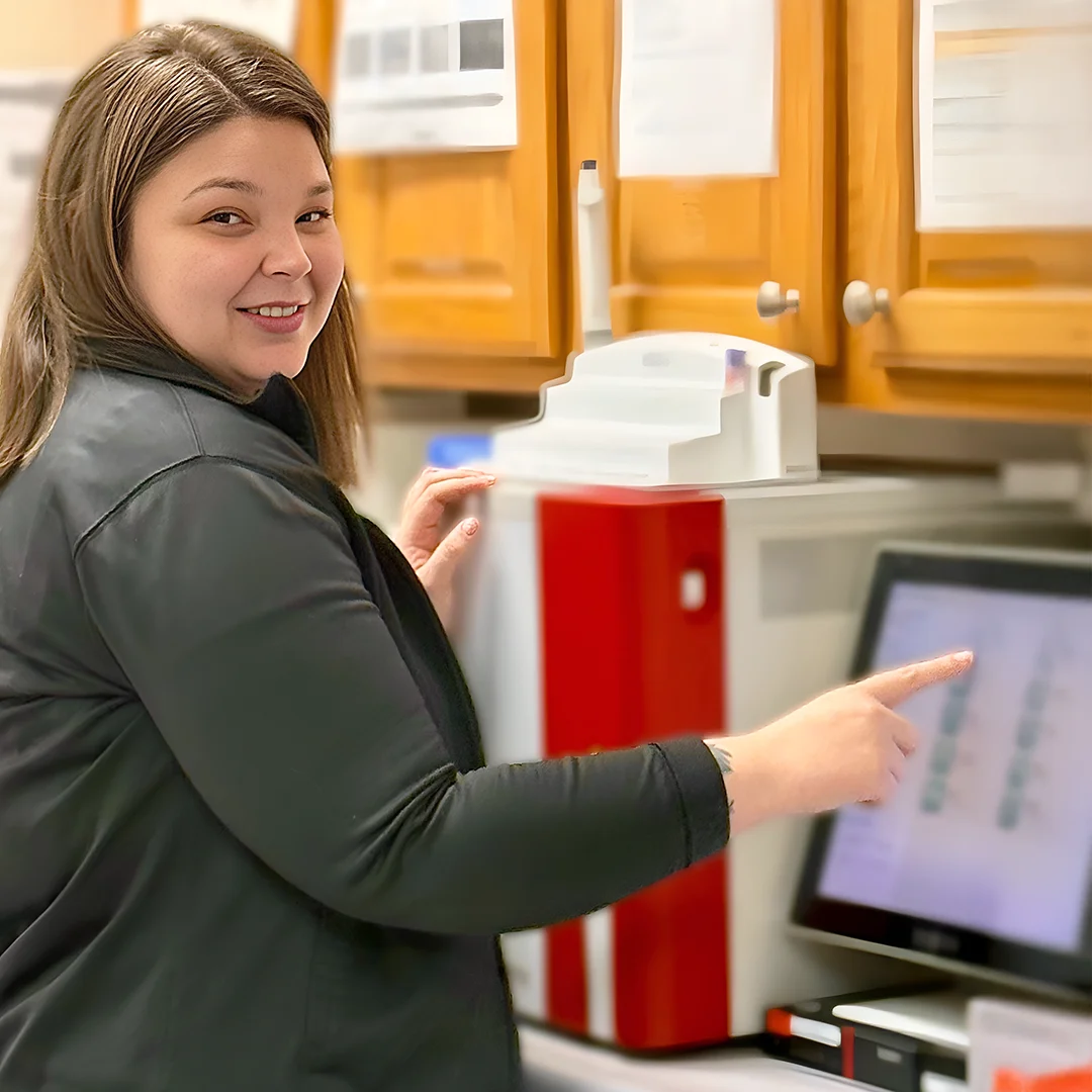 Ashley Dorador – Woman in a black jacket smiling while using a touchscreen device in a medical or lab setting.