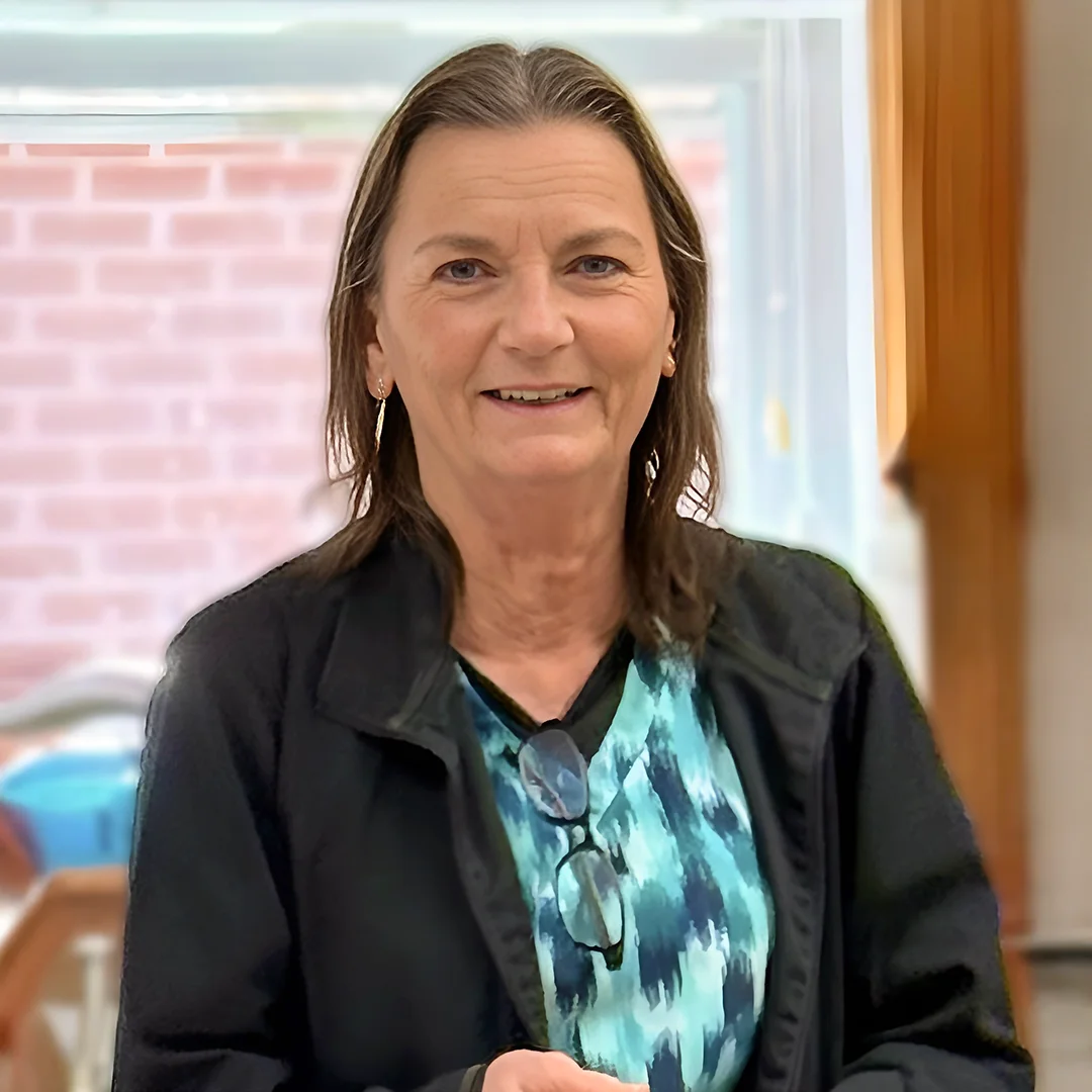 Debbie Kittrell – Woman in a patterned shirt and black jacket smiling indoors near a brick wall and window.