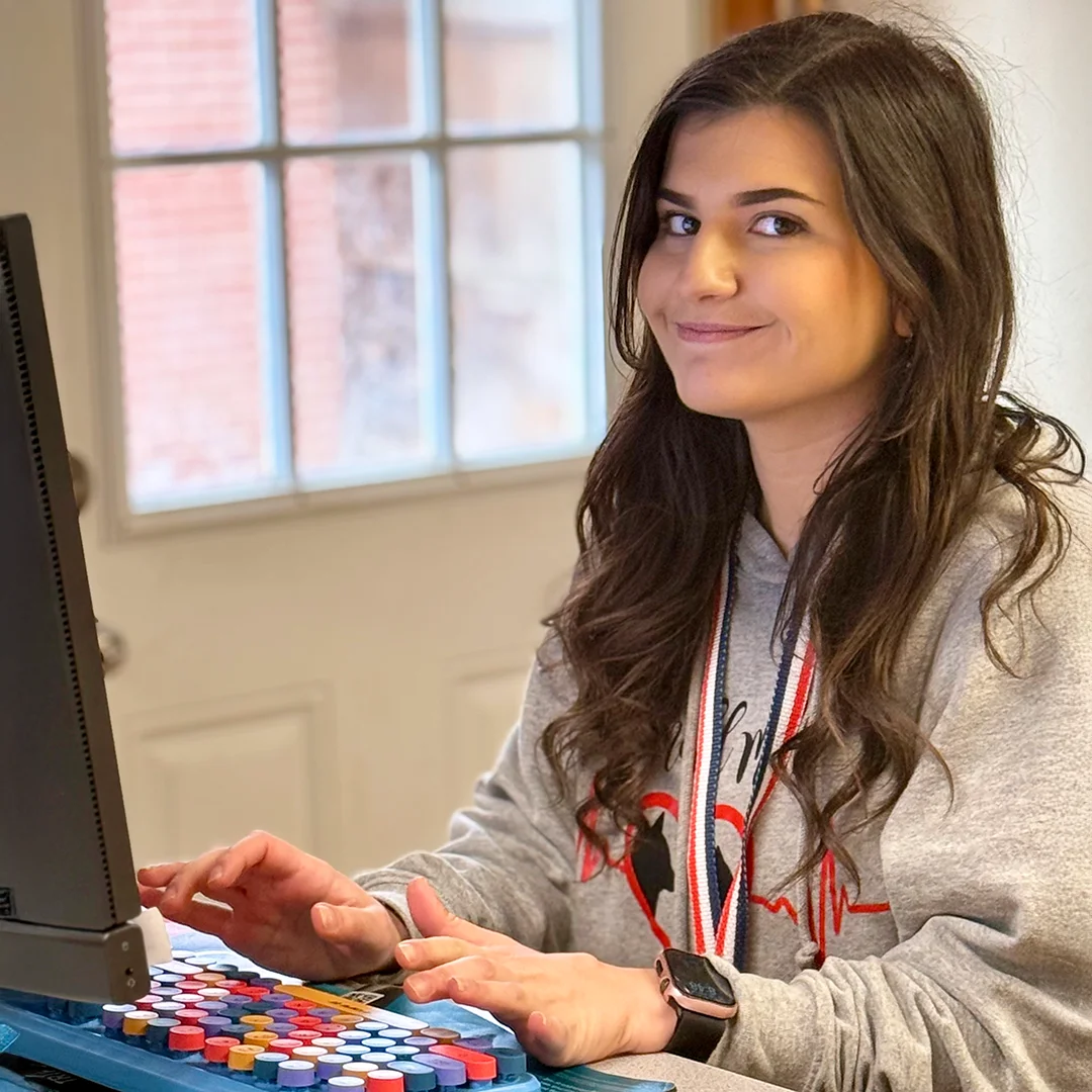 Leanna Frye – Woman in a gray hoodie smiling while working on a computer with colorful keys.
