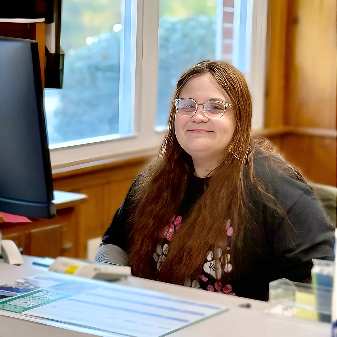 Morgan Phillips – Woman with long brown hair and glasses sitting at a reception desk and smiling.