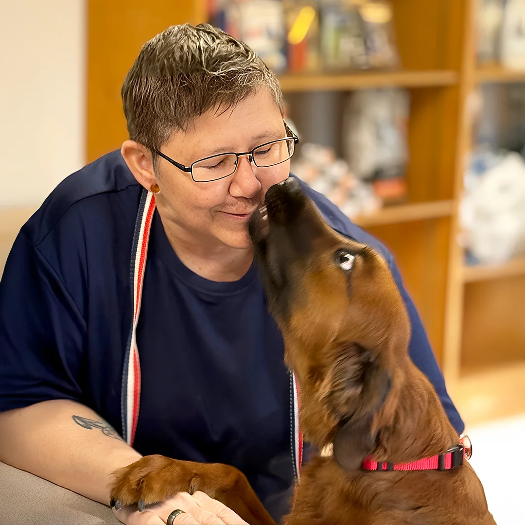 Stephanie Adams – Person with short hair and glasses smiling while being licked on the face by a brown dog.